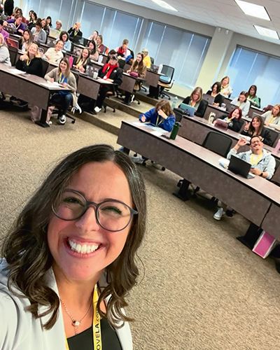 Kate is smiling while taking a selfie with her classroom of students in the background