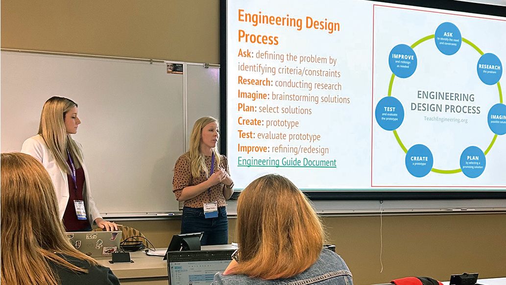 Maggie and Kayla are having a presentation to their class. A slide is on the projector screen behind them with the main header, Engineering Design Process.