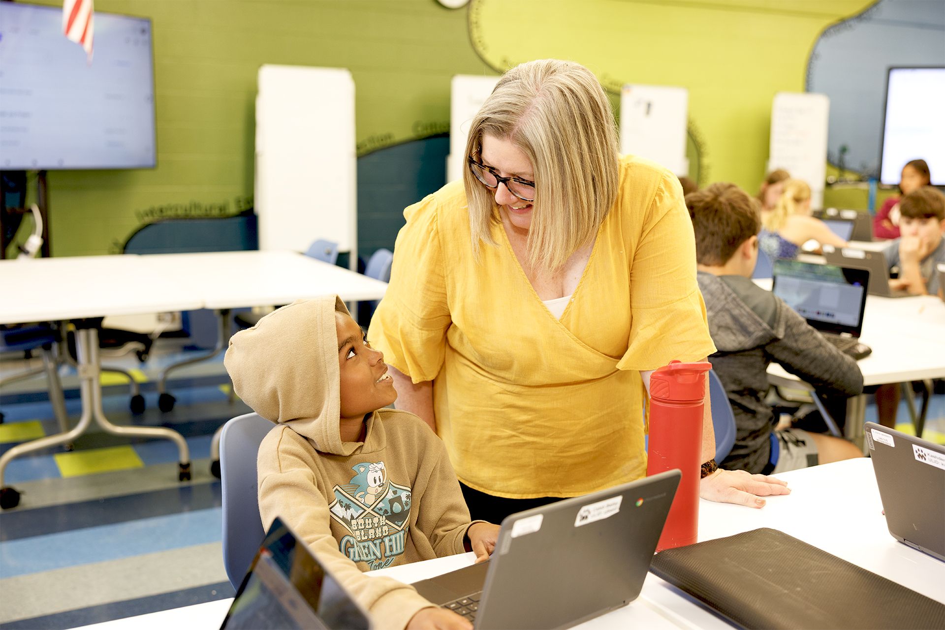 Kristi Sutter, wearing a yellow top, leans down to smile and converse with student Caleb Bailey, who looks up warmly from his laptop at a classroom table.