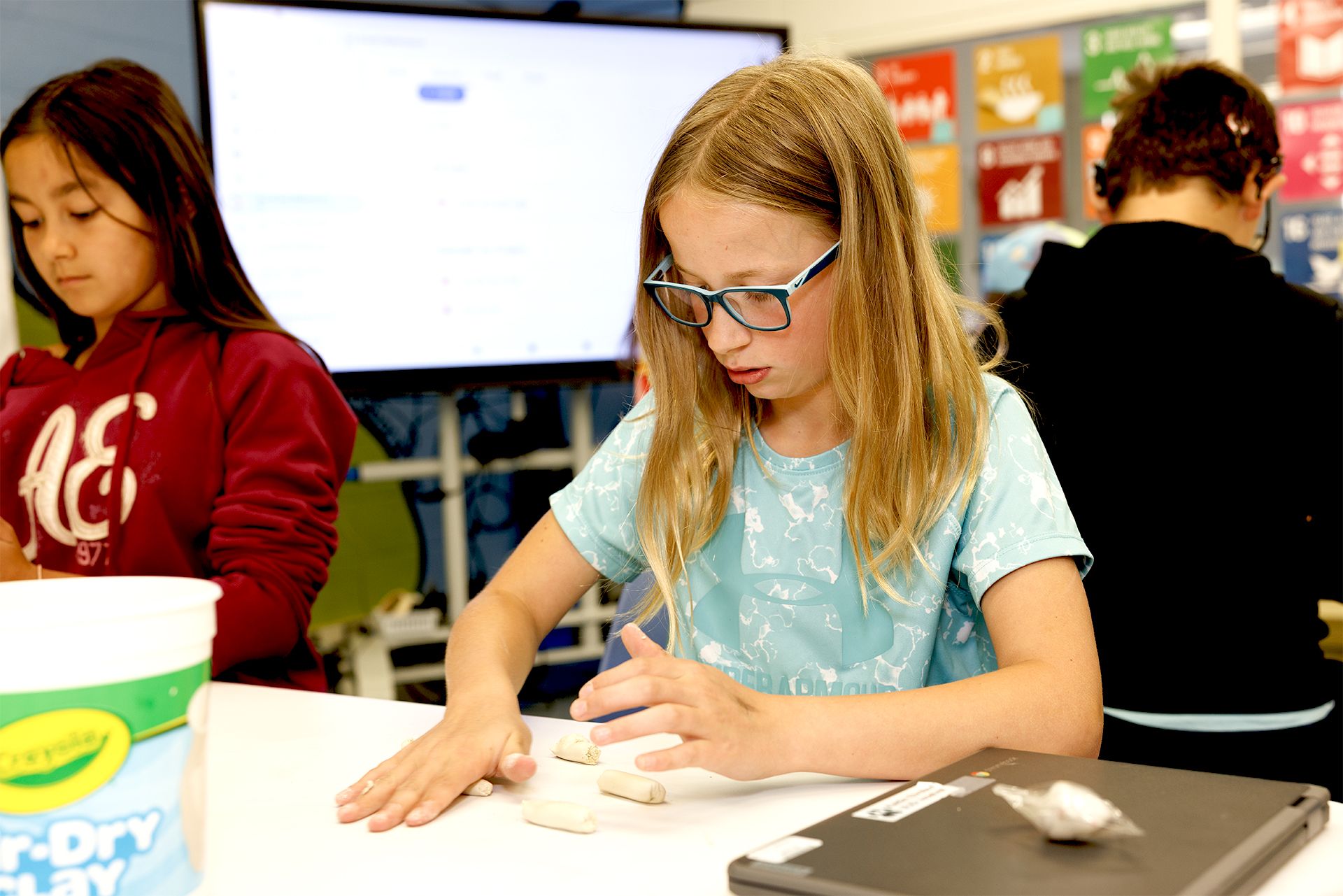 Student Kaitlin Ehrstein wears blue-framed glasses and a light blue t-shirt as she intently shapes small pieces of clay on a table in a classroom setting.