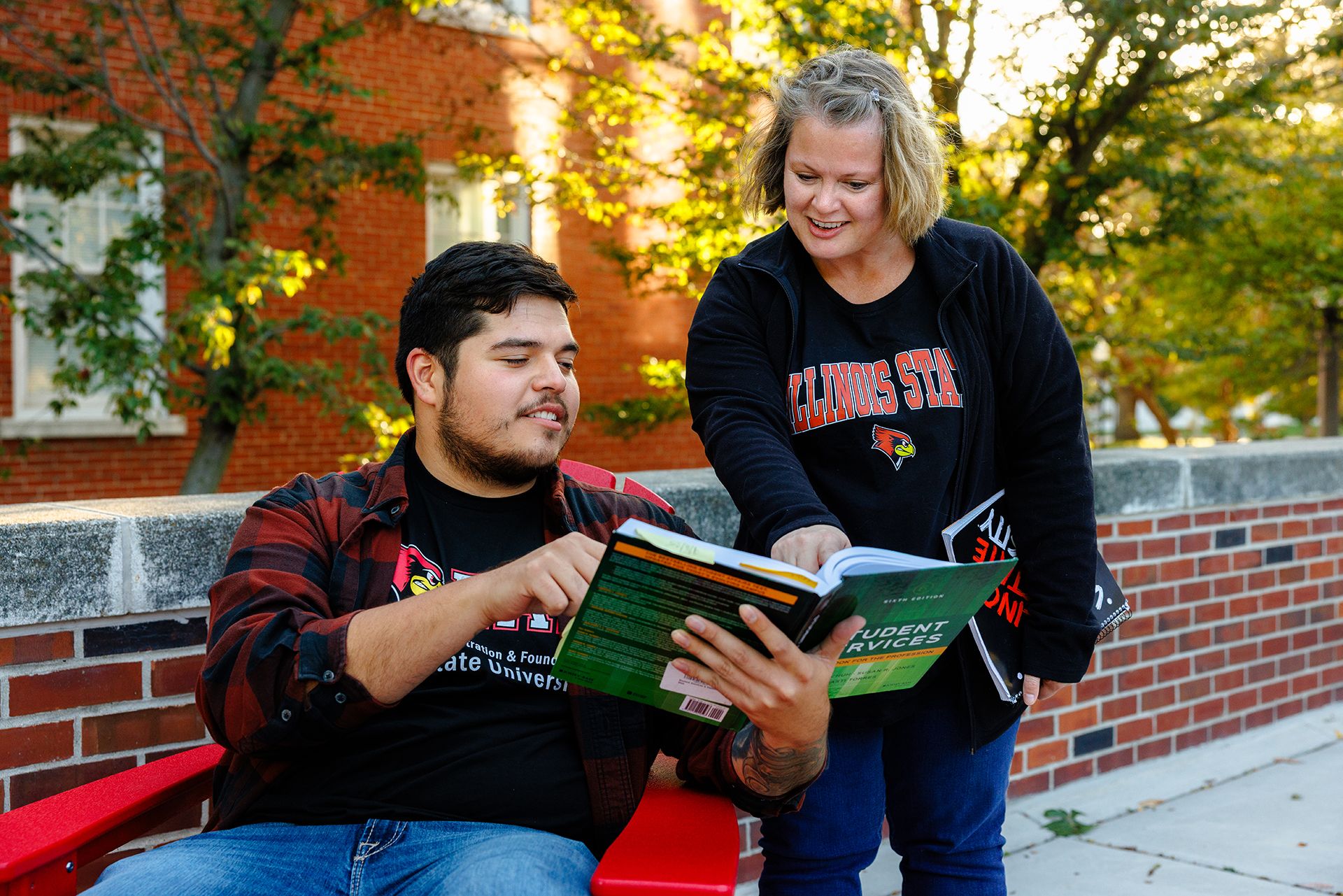 Two university students read an open book together outdoors, with a brick building and trees behind them; the book says “STUDENT SERVICES.”