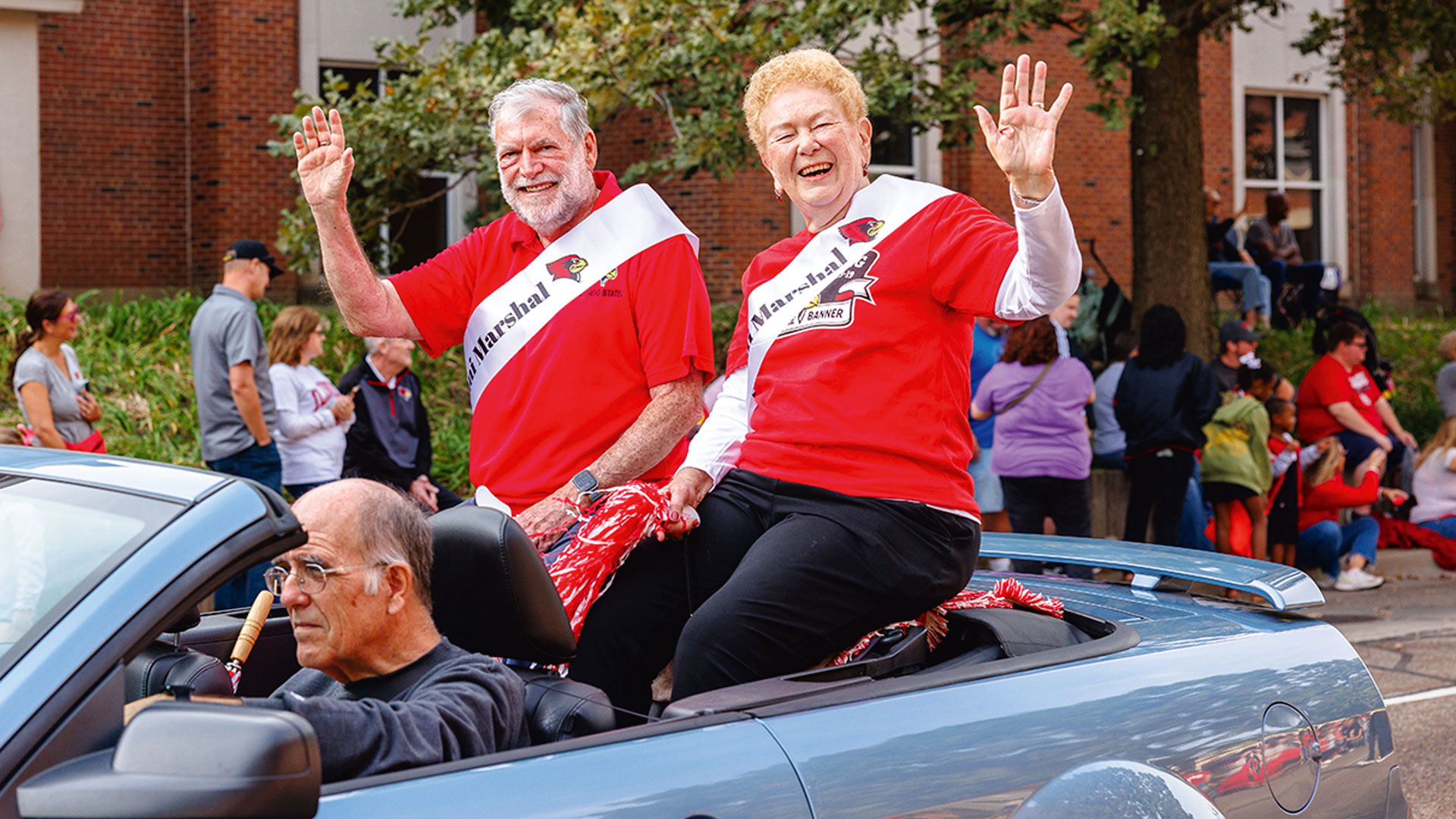 Homecoming 2025 alumni marshals Mike Schermer and Glenna Adams wave to a parade crowd while riding in the back of a light blue convertible.