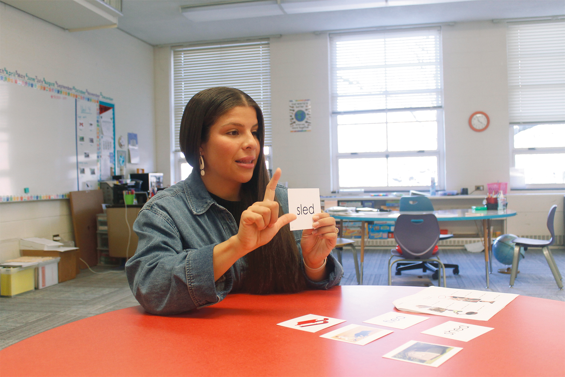 An individual holds up a card labeled “sled” while pointing to it, with picture cards spread across a classroom table.