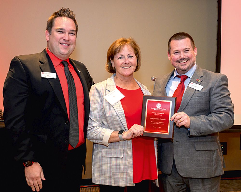 Three people pose indoors, holding a red plaque that reads “Chamber of Commerce.”