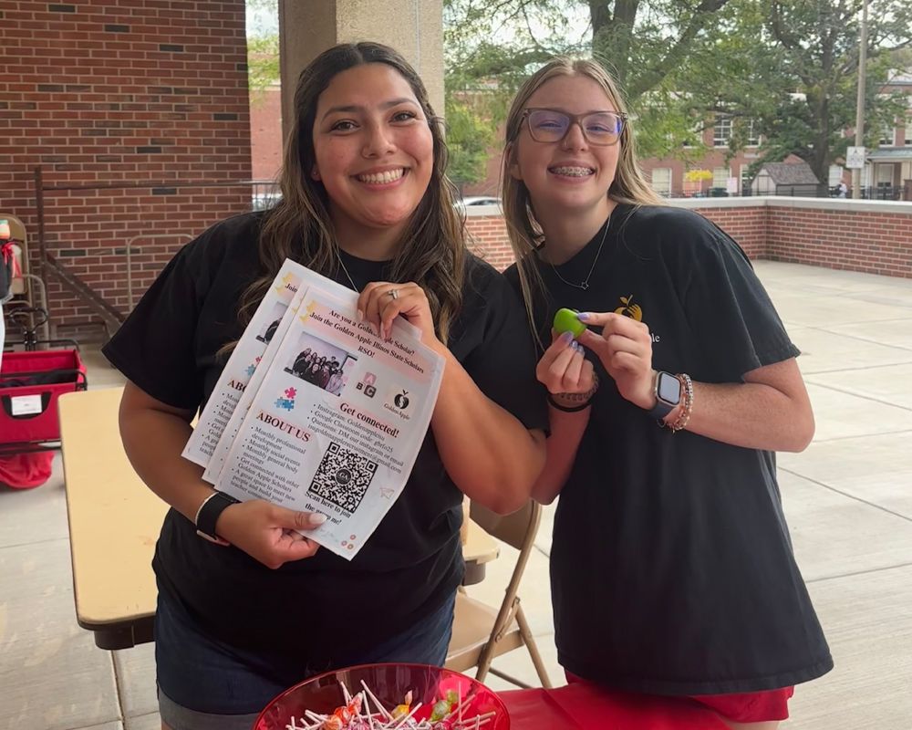 Two smiling students stand at a table; one holds a flyer with a QR code, while the other holds a small green object.