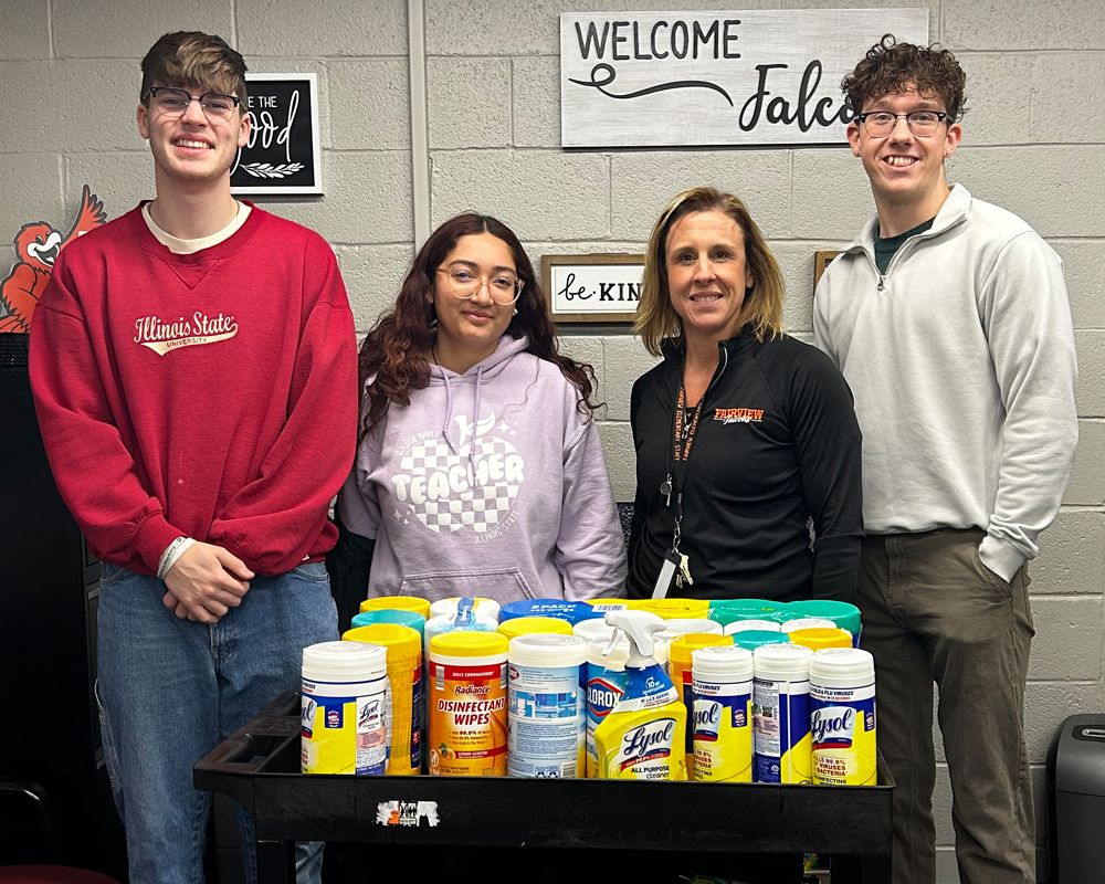 Four people stand behind a cart filled with disinfectant wipes and cleaning products; signs behind them read “Welcome” and “be kind.”
