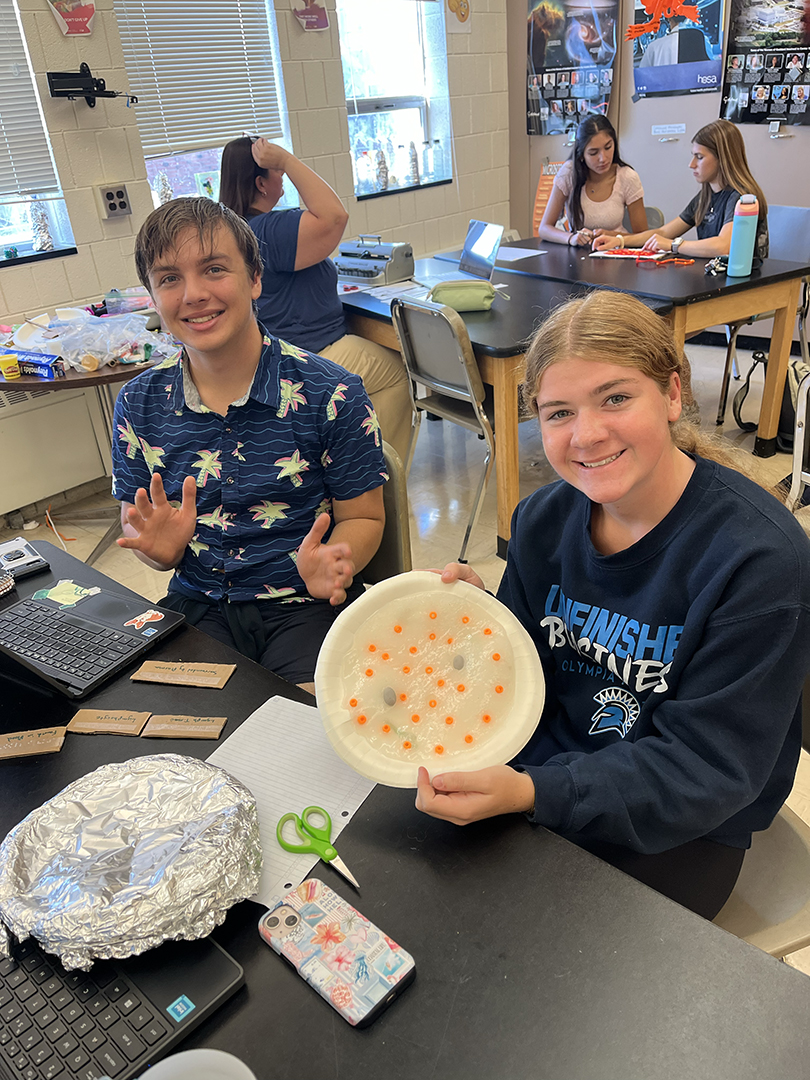 Two students sit at a classroom table; one holds a plate of white material with orange and gray dots while the other gestures.