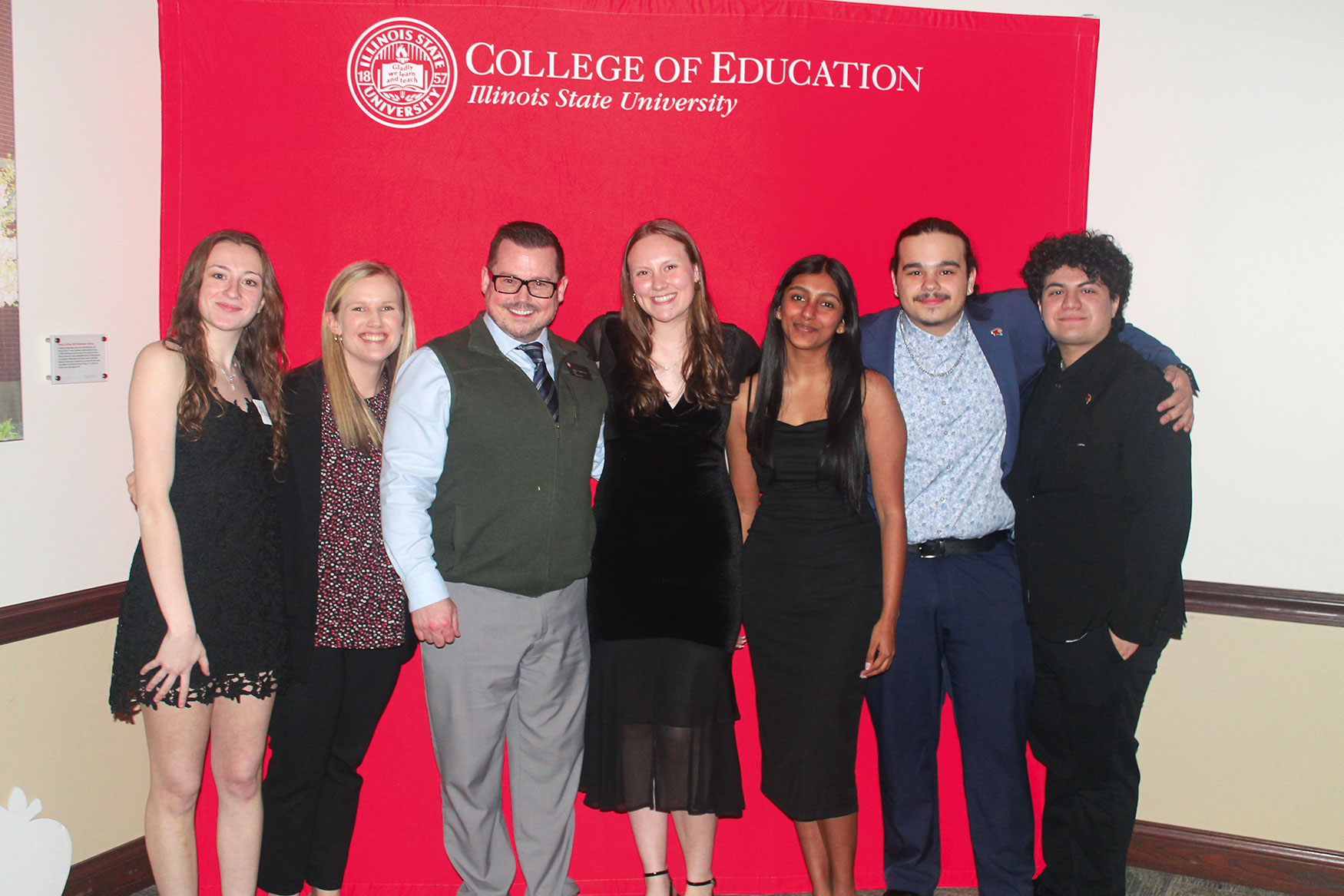 The seven members of the Inaugural Executive Leadership Team standing together and smiling in front of a red Illinois State University College of Education banner.