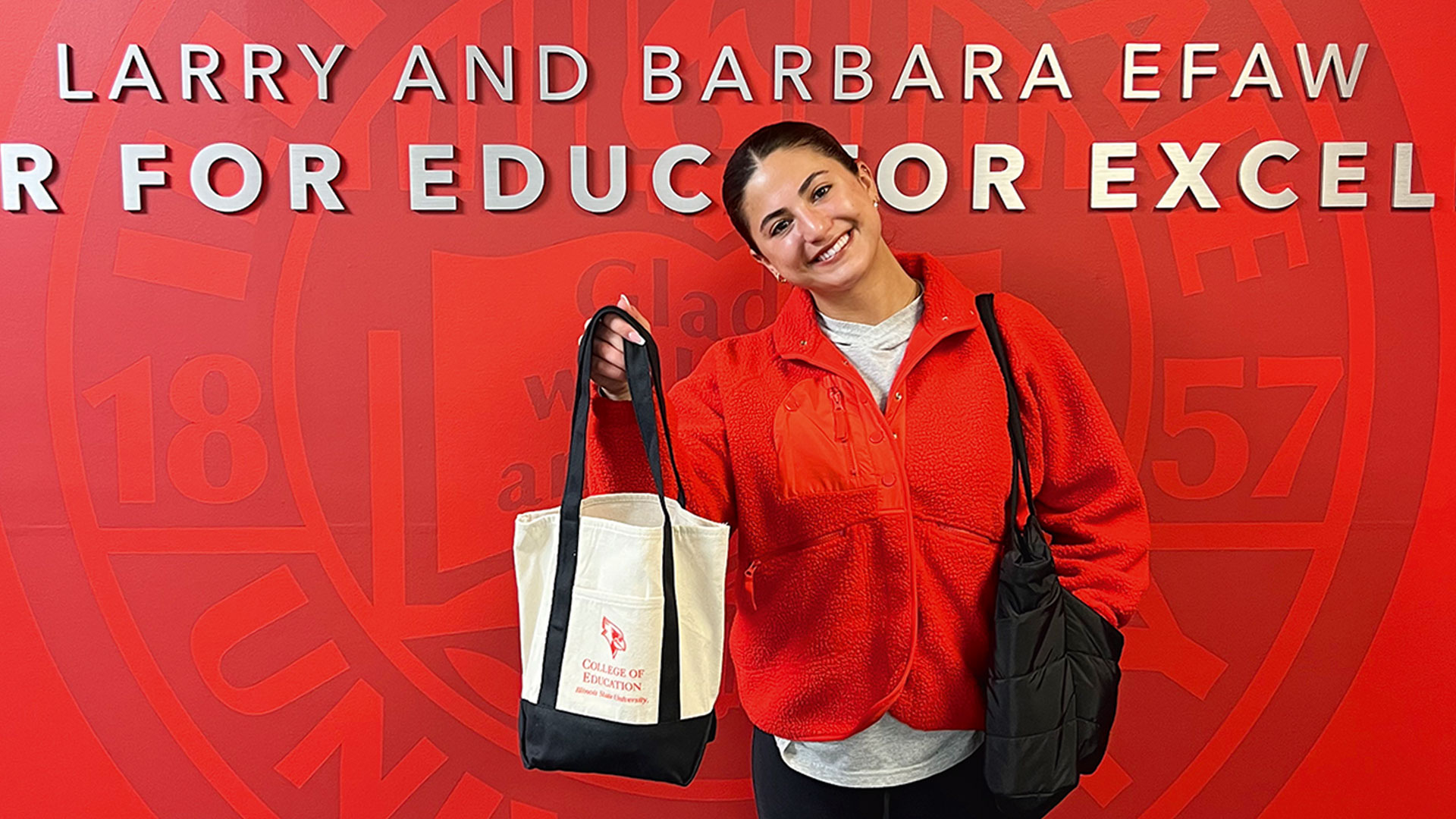 Zoe Limparis, wearing a bright red fleece jacket, smiles broadly as she holds up a canvas College of Education tote bag in front of a red wall that reads Larry and Barbara Efaw center for educator excellence.