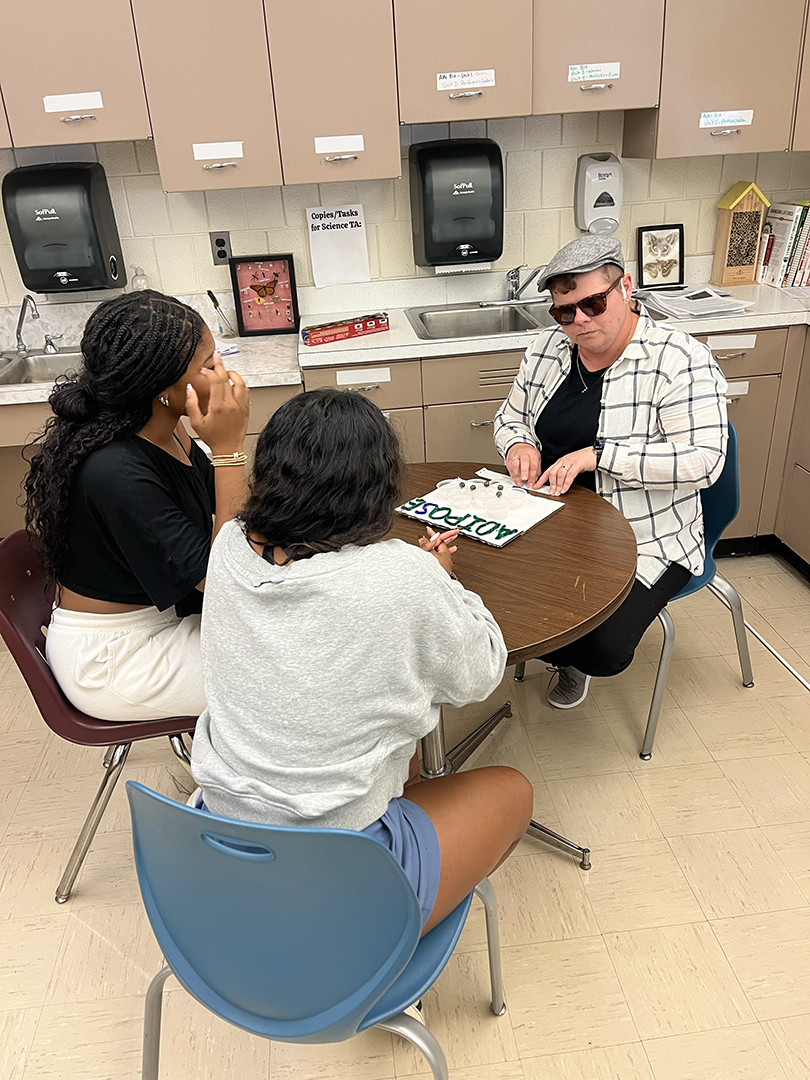 Dr. Shaheen, wearing sunglasses and a newsboy cap, sits at a round table reading Braille while two students look on. On the table is a tactile educational model labeled "ADIPOSE" with raised text and shapes.
